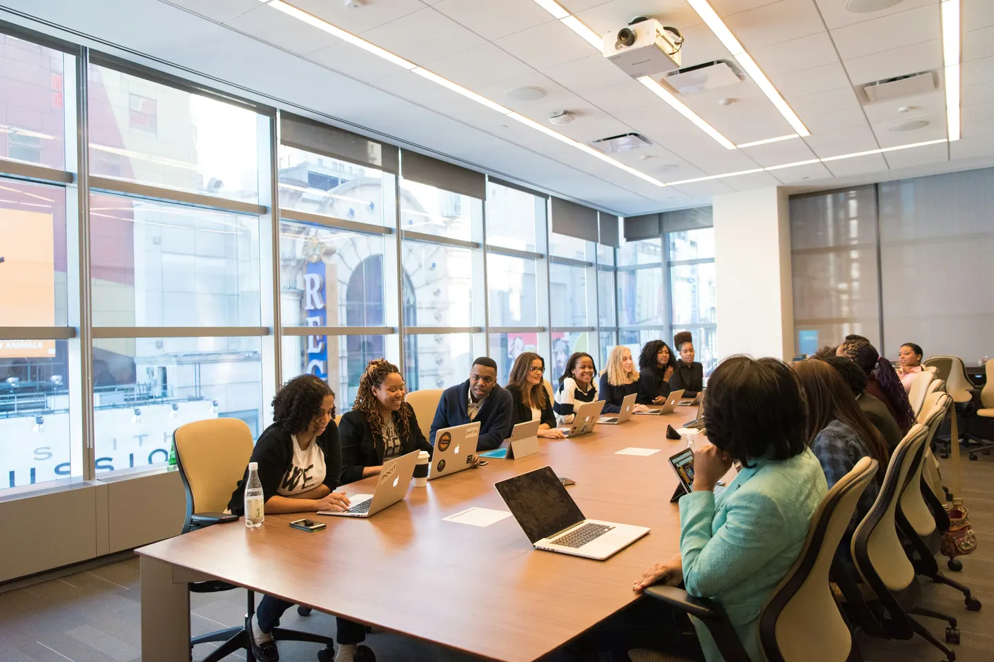 A group of people seated along a long table with laptops in a bright room with tall windows